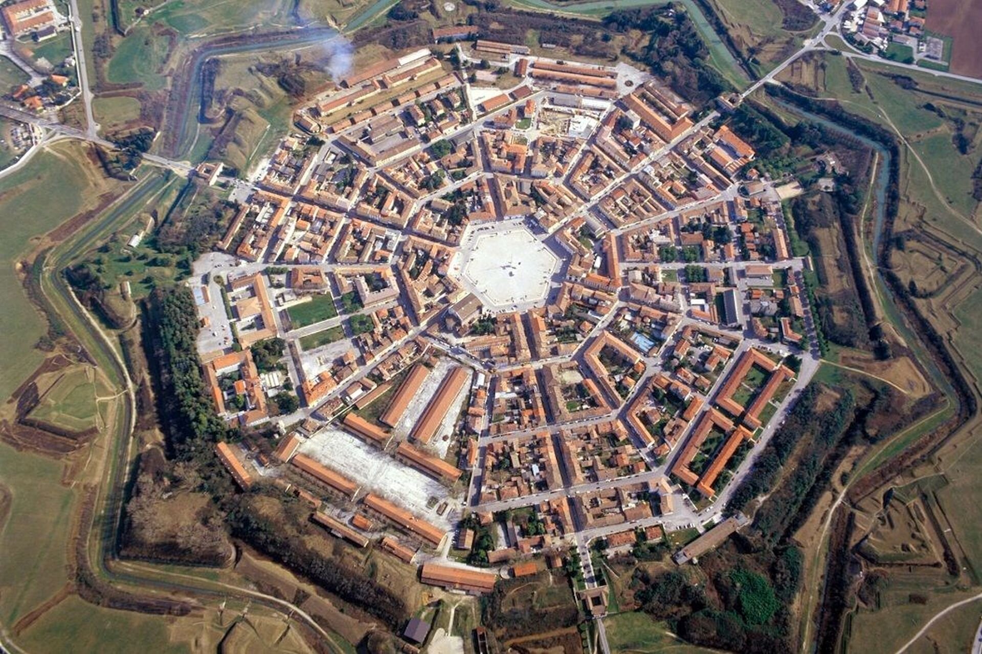 Ilustracja przedstawia zdjęcie miasta Palmanova z lotu ptaka. Ukazuje założenie w kształcie dziewięcioramiennej gwiazdy. Miasto otaczały bastiony, mury, fosa i trzy bramy. Z centrum znajduje się sześciokątny rynek, od którego rozchodzą się promieniście ulice.