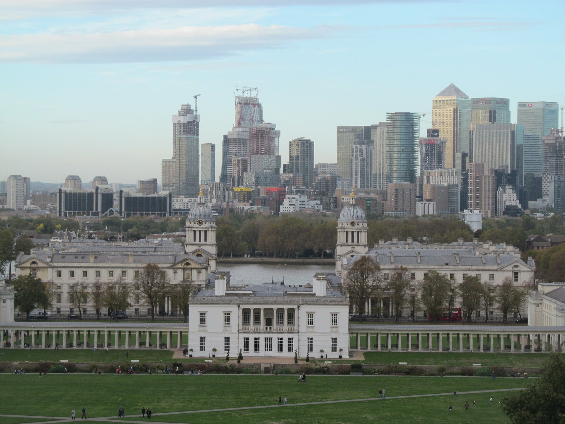 Zdjęcie przedstawia Greenwich Park w Londynie. Fotografia ukazuje panoramę miasta. Na pierwszym planie ukazany jest biały budynek w kształcie sześcianu. Na środku budynku znajduje się taras, który ma cztery kolumny. Budynek ma prostokątne okna. Po prawej oraz po lewej stronie znajdują się dwa zadaszone skrzydła z kolumnami. Budynek usytuowany jest w parku, w którym spacerują różne postacie. Na drugim planie przedstawiony jest budynek z dwoma kopułami, na których znajdują się czarne zegary ze złotymi wskazówkami. W oddali znajduje się rzeka oraz dalej panorama miasta, gdzie znajdują się liczne drapacze chmur.