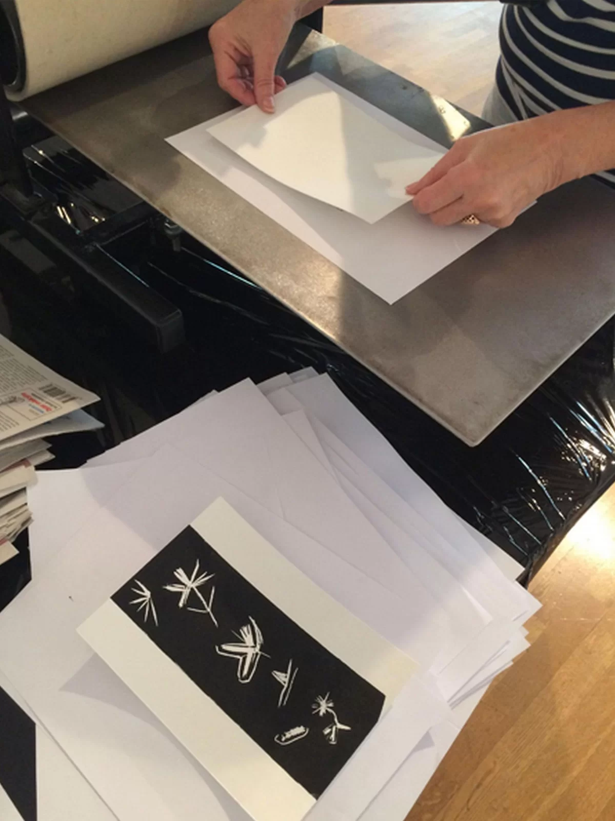 Ilustracja przedstawia fragment postaci z dłońmi, trzymającymi w palcach białą kartkę na matrycy. U dołu leży gotowa odbitka z motywami kwiatowymi na czarnym tle, położona na stosie białych kartek.