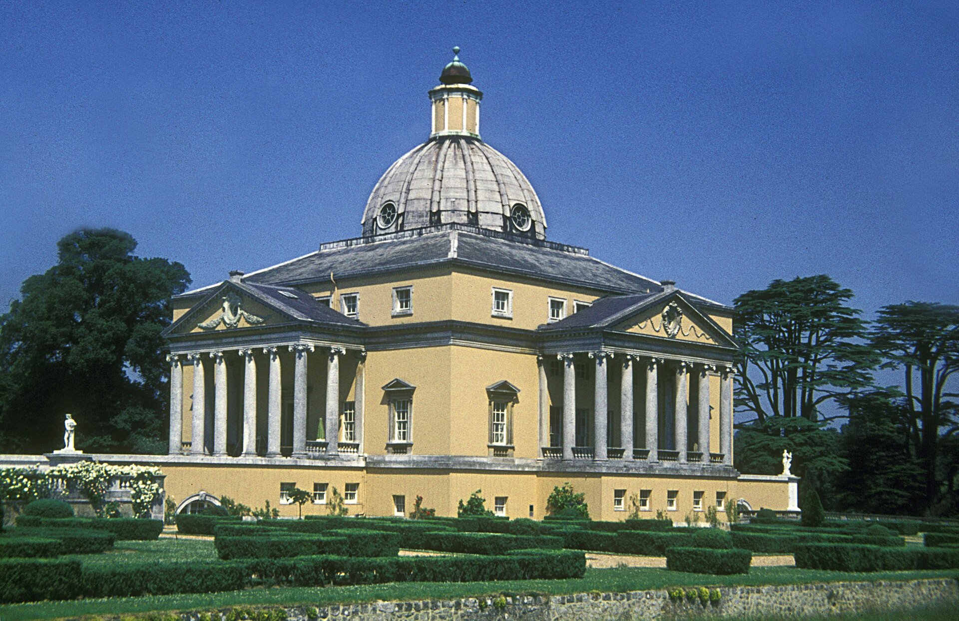 Zdjęcie przedstawia Zamek Mereworth w Wielkiej Brytanii. Fotografia przedstawia żółty zamek, znajdujący się w ogrodzie. Z lewej oraz prawej strony zamku znajdują się tarasy otoczone szarymi kolumnami. Nad tarasami znajduje się tympanon z ozdobną płaskorzeźbą. Dach tworzy szara kopuła z dwoma okrągłymi oknami. W budynku znajdują się białe okna: większe- prostokątne oraz mniejsze- kwadratowe. Cały kompleks usytuowany jest wśród drzew oraz krzewów. Po lewej stronie znajdują się krzew z białymi kwiatami.