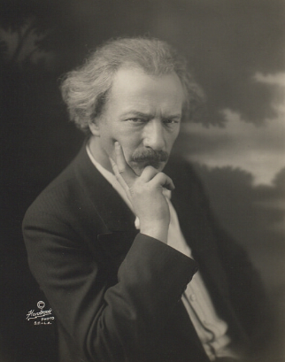 Ilustracja przedstawia portret mężczyzny w średnim wieku. Ma on siwe włosy do ramion, mocne rysy twarzy, wydatny nos oraz wąsy. Ubrany jest w garnitur.