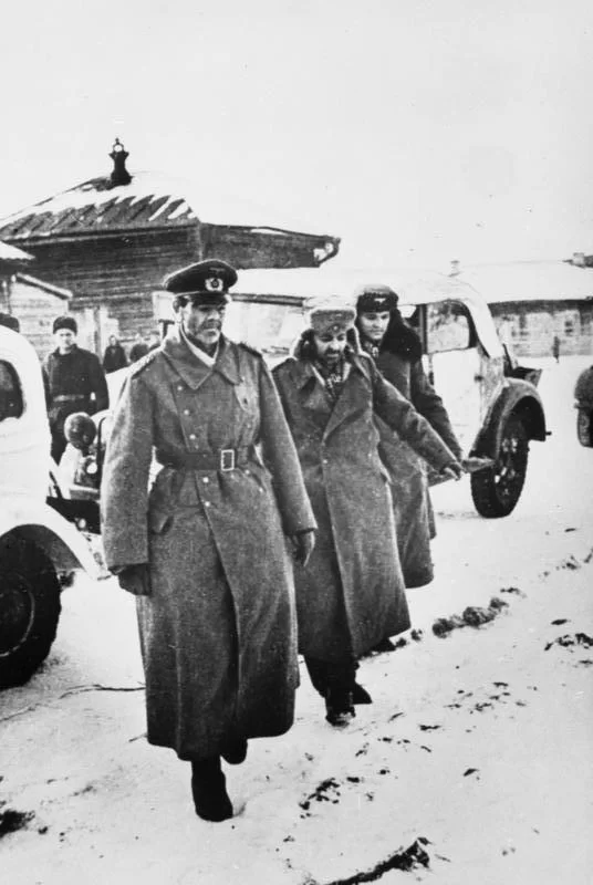 Kliknij, aby powiększyć Na zdjęciu niemiecki oficer w wojskowym płaszczu idzie w asyście dwóch sowieckich żołnierzy. W tle widać dwa samochody i wiejskie zabudowania. Na ziemi leży śnieg.
