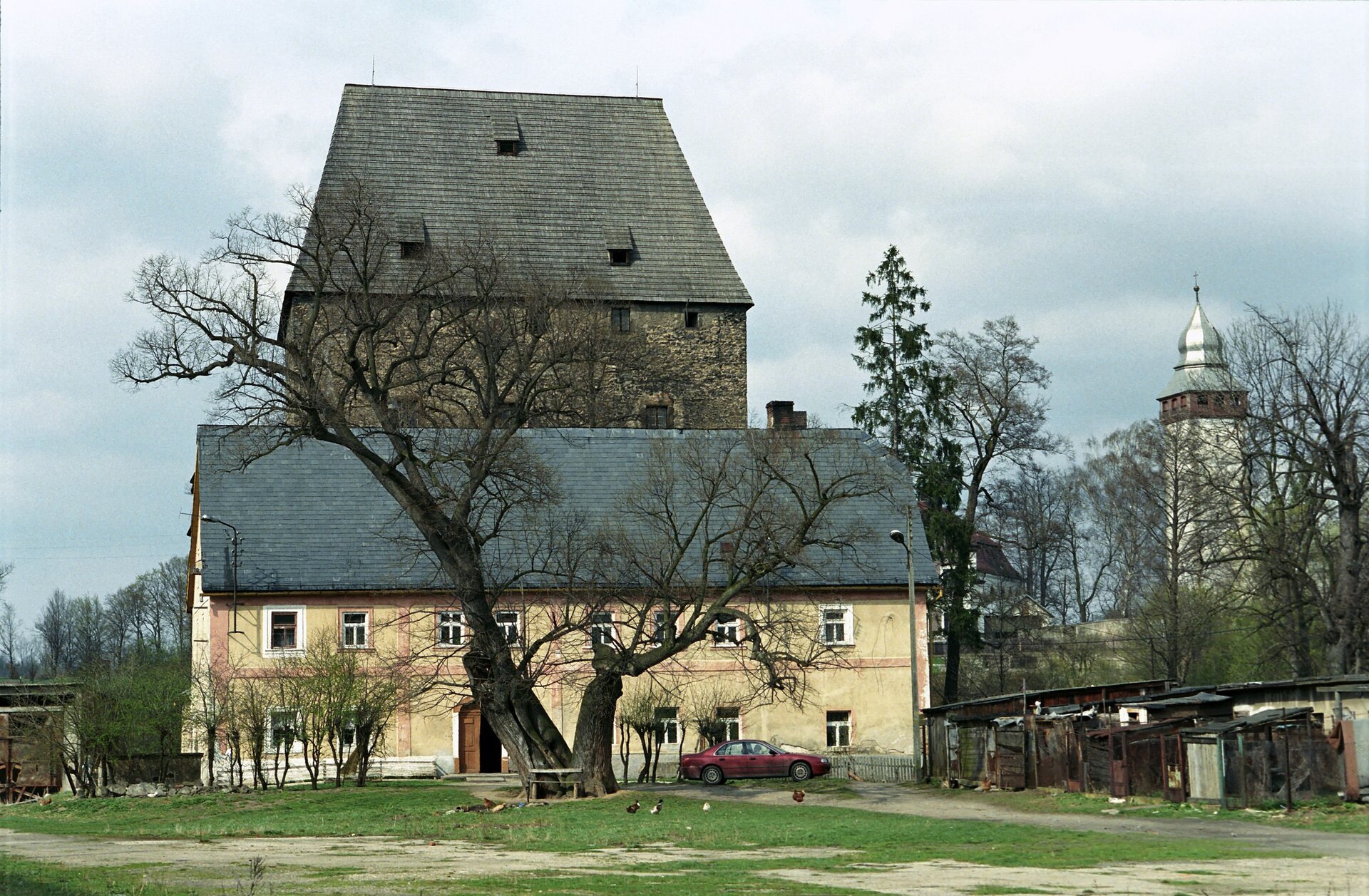 Fotografia przedstawia współczesny widok na wieżę we wsi Siedlęcin. Na zdjęciu widoczny jest piętrowy budynek, który dobudowany został do wieży. Budynek ma skośny dach i żółtą elewację. Za nim znajduje się kilkupiętrowa wieża, której górne kondygnacje widoczne są na tle nieba. Wieża jest wykonana z ciemnobrązowych cegieł i ma skośny dach. Wokół widać drzewa, a przed budynkiem zaparkowany jest samochód osobowy. Z prawej strony zdjęcia widoczne są niskie zabudowania mieszkalne, a także wieża ze srebrnym, stożkowym dachem, na czubku którego znajduje się krzyż.