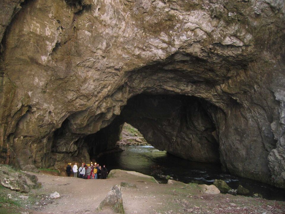 Fotografia przedstawia ponor - otwór szczelinowy wydrążony przez wodę. Jest to duża dziura w skale, do której wpływa woda. Na tle skały stoi kilkunastoosobowa grupa ludzi. 