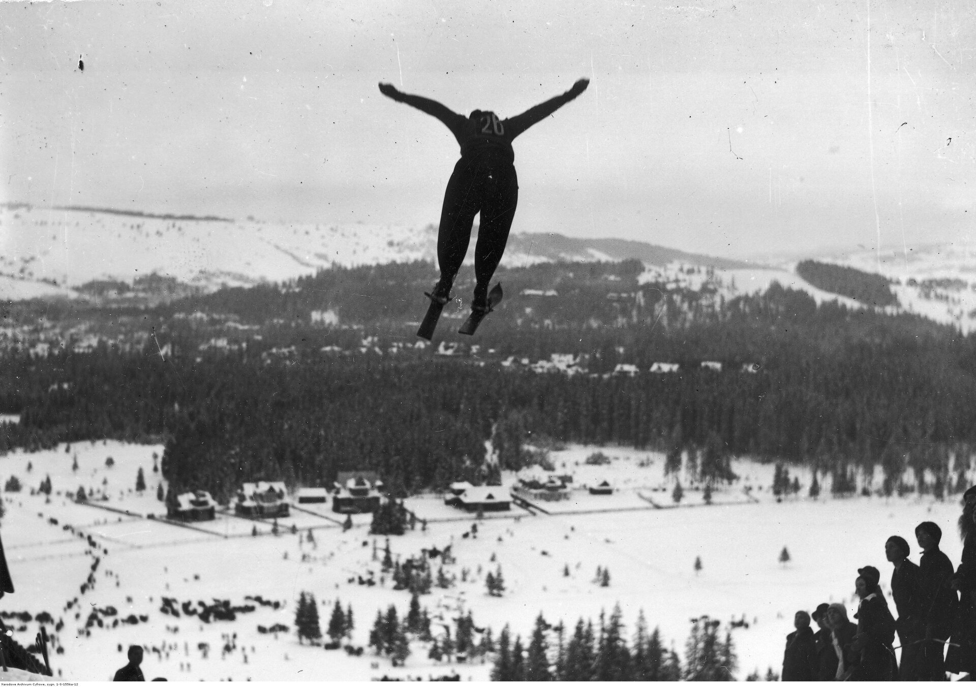 Czarno - białe zdjęcie skoczka narciarskiego, lecącego nad zboczem góry, po wykonaniu skoku. 
Z prawej strony u dołu zdjęcia widnieje grupa  kibiców.  