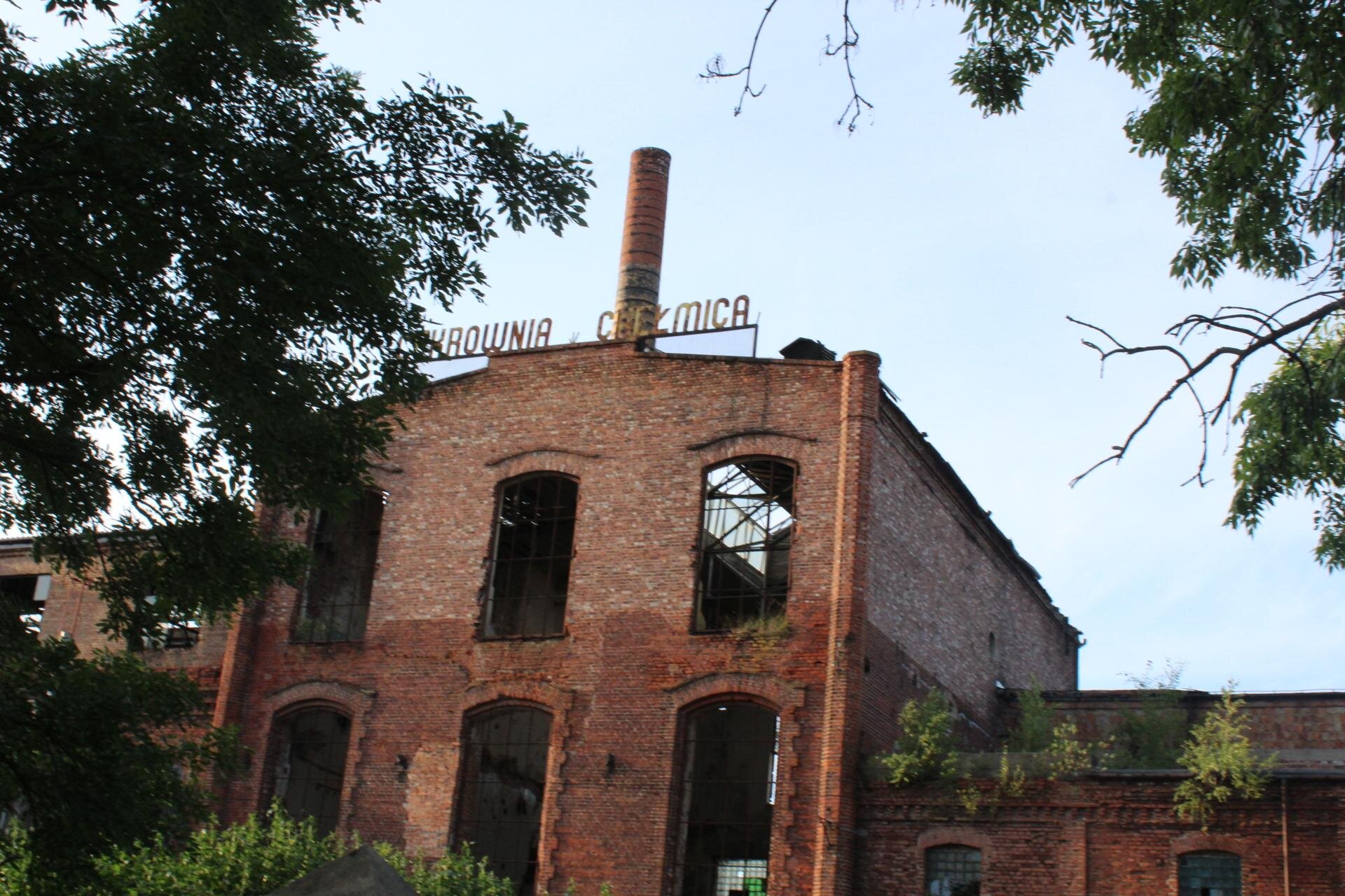 Zdjęcie przedstawia piętrowy, ceglany budynek. Miejsca na okna są puste, bez szyb. Na dachu budynku znajduje się napis "CUKROWNIA CHEŁMICA". W tle widoczny jest wysoki, pomarańczowy komin. Po prawej i lewej stronie zdjęcia widoczne są fragmenty zielonych, wysokich drzew. Niebo jest błękitne, bezchmurne. Zdjęcie zostało wykonane w świetle dziennym.