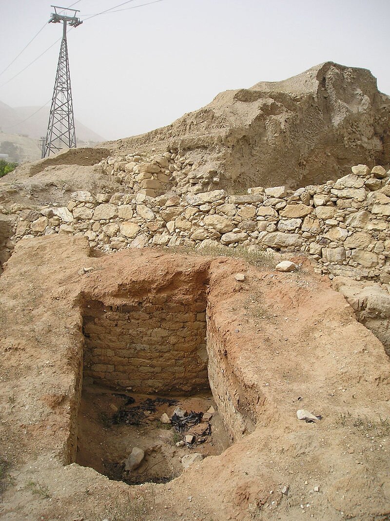 Zdjęcie przedstawia fragment wykopalisk. Fragmenty muru z jasnych kamieni. Przed nim otwór w ziemi, wzmocniony glinianymi cegłami i od góry obłożony gliną. Dalej kawałek ruin piaskowych. W tle współczesny słup trakcji elektryczny.  