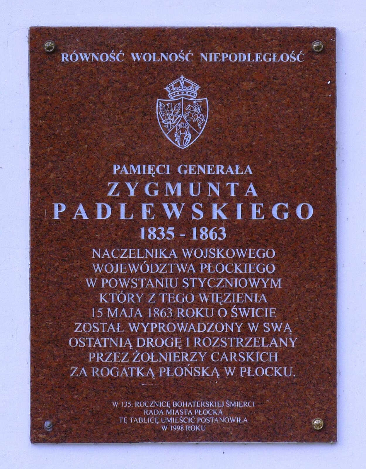 Zdjęcie przedstawia brązową tablicę pamiątkową z wyrytymi na niej białymi napisami. Tablica wisi na białej ścianie. Na tablicy pojawiają się napisy: "RÓWNOŚĆ, WOLNOŚĆ, NIEPODLEGŁOŚĆ". Poniżej znajduje się herb podzielony na trzy części - w jednej z nich pojawia się orzeł, w drugim anioł z mieczem, a w trzecim rycerz na koniu. Herb u góry ma nałożoną czapkę. Poniżej znajdują się napisy: "PAMIĘCI GENERAŁA ZYGMUNTA PADLEWSKIEGO, 1835 - 1863. NACZELNIKA WOJSKOWEGO WOJEWÓDZTWA PŁOCKIEGO W POWSTANIU STYCZNIOWYM, KTÓRY Z TEGO WIĘZIENIA 15 MAJA 1863 ROKU O ŚWICIE ZOSTAŁ WYPROWADZONY W SWĄ OSTATNIĄ DROGĘ I ROZSTRZELANY PRZEZ ŻOŁNIERZY CARSKICH ZA ROGATKĄ PŁOŃSKĄ W PŁOCKU. W STO TRZYDZIESTĄ PIĄTĄ ROCZNICĘ BOHATERSKIEJ ŚMIERCI RADA MIASTA PŁOCKA TĘ TABLICĘ UMIEŚCIĆ POSTANOWIŁA W 1998 ROKU".