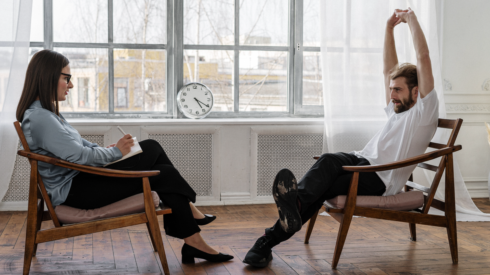 Na zdjęciu dwie osoby siedzą na krzesłach na przeciwko siebie. Kobieta po lewej stronie jest bardzo elegancko ubrana ma okulary. Na kolanie trzyma zeszyt, w którym notuje. Po prawej stronie, na przeciwko kobiety siedzi młody mężczyzna. Ręce ze splecionymi palcami ma wyciągnięte ku górze, wygląda jakby się przeciągał. W tle, na parapecie znajduje się zegar wskazujący godzinę piątą dwadzieścia.