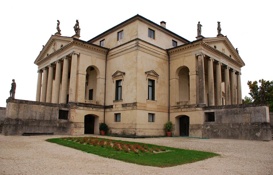 Zdjęcie przedstawia budynek dwukondygnacyjny. Z prawej i z lewej strony budowli znajdują się portyki zwieńczone frontonem. Na ich krawędziach stoją  po dwie rzeźby. Przed budynkiem znajduje się trawiasty prostokąt, porośnięty kwiatami na środku. Po prawej stronie budowli rośnie drzewo.