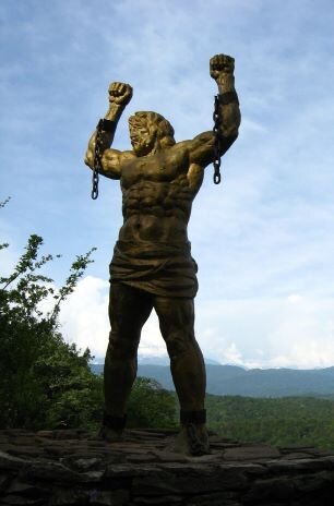Fotografia przedstawia monumentalna rzeźbę mężczyzny stojącą w plenerze. Mężczyzna jest prawie nagi, jedynie  niewielka szata osłania jego biodra. Jest umięśniony. Zaciśnięte w pięści ręce, z zerwanymi kajdanami, unosi do góry. Stopy wtopione ma w skałę, na której stoi. W tle znajdują się wierzchołki drzew oraz szczyty gór. 