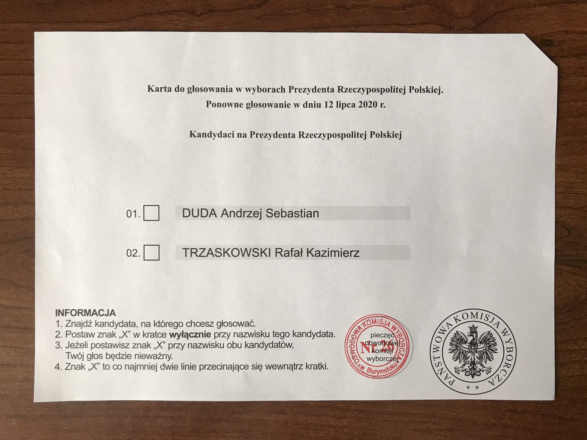 Na zdjęciu widoczna karta do głosowania z napisem: Karta do głosowania w wyborach Prezydenta Rzeczypospolitej Polskiej. Ponowne głosowanie w dniu 12 lipca 2020 r. Kandydaci na Prezydenta Rzeczypospolitej Polskiej. Podane są dwa nazwiska: Duda Andrzej Sebastian i Trzaskowski Rafał Kazimierz, po lewej stronie nazwisk znajdują się pola do zaznaczenia X. Poniżej informacja: 1. Znajdź kandydata, na którego chcesz głosować. 2. Postaw znak "X" w kratce wyłącznie przy nazwisku tego kandydata. 3. Jeżeli postawisz znak "X" przy nazwisku obu kandydatów, Twój głos będzie nieważny. 4. Znak "X" to co najmniej dwie linie przecinające się wewnątrz kratki. U dołu kartki znajduje się pieczęć Obwodowej Komisji Wyborczej i pieczęć Państwowej Komisji Wyborczej.Pieczęć Obwodowej Komisji Wyborczej w Białymstoku nr 26 jest okrągła. Napis  w kolorze czerwonym.Pieczęć Państwowej Komisji Wyborczej jest okrągła. W kolorze czarnym. Z godłem Rzeczypospolitej Polskiej.