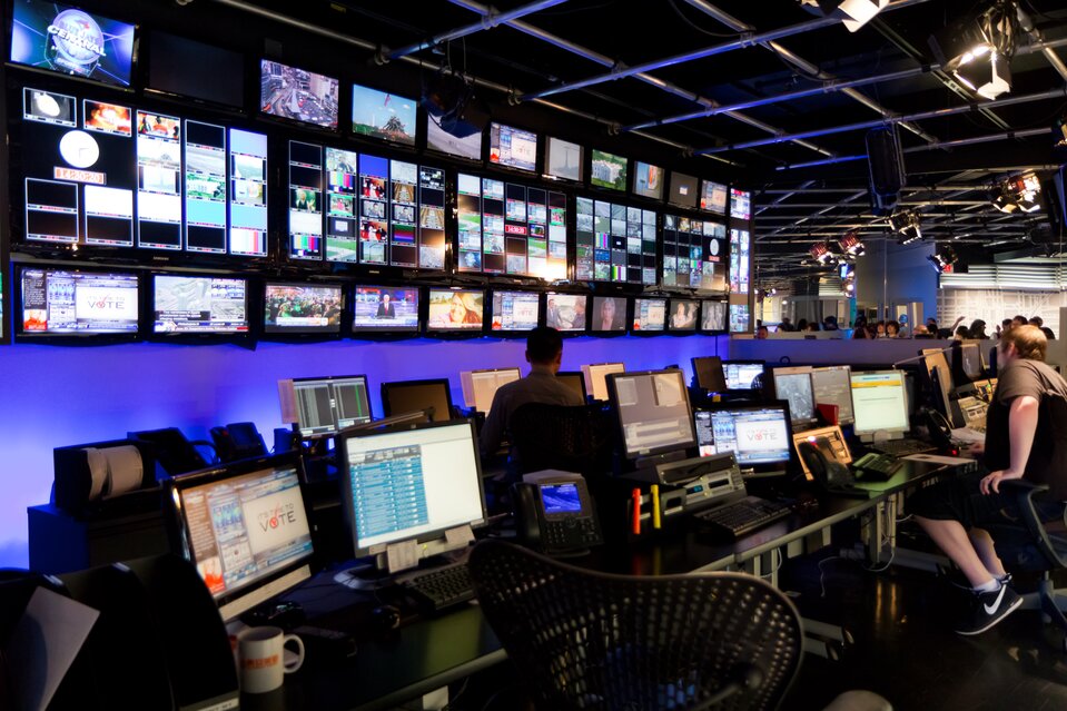 Na zdjęciu pomieszczenie tzw. reżyserki w telewizji w Kanadzie: U góry od połowy ściany aż do sufitu umieszczone są monitory z podglądem na transmisję z różnych kamer. W pomieszczeniu stoją dwa długie rzędy stołów, na których znajdują się włączone komputery. W jednym rzędzie przy jednym z komputerów siedzi pracownik reżyserki, w drugim rzędzie - inny.  Na suficie są umieszczone reflektory ze strumieniem światła skierowanym na miejsca siedzące. Na dalszym planie znajdują się kolejne stanowiska komputerowe i dość duża grupa pracowników.