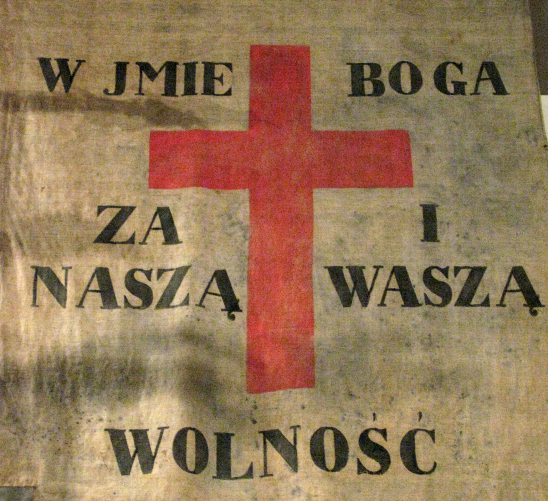 Zdjęcie przedstawia stary, przybrudzony sztandar, na którym znajduje się duży, czerwony krzyż oraz czarny napis: W imię Boga za naszą i waszą wolność.