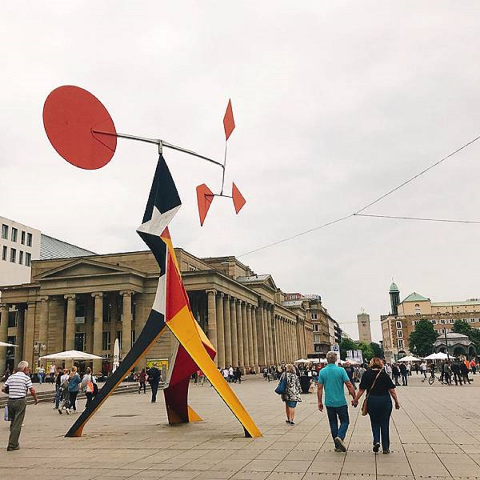 Fotografia przedstawia rzeźbę wykonaną z kolorowej blachy - jej kolory to żółty, biały, czerwony i czarny.. Przedstawia abstrakcyjny, geometryczny kształt. Ma trzy nogi, które później zwężają się w jedną całość, a na szczycie umieszczono poziomy pręt. Na jednym końcu pręta jest zamocowane czerwone koło z blachy, a na drugim - trzy wieloboczne kształty w kolorze czerwonym, połączone prętami. Rzeźba stoi na deptaku, po którym chodzi wiele ludzi. W tle stoją budynki i rosną drzewa.