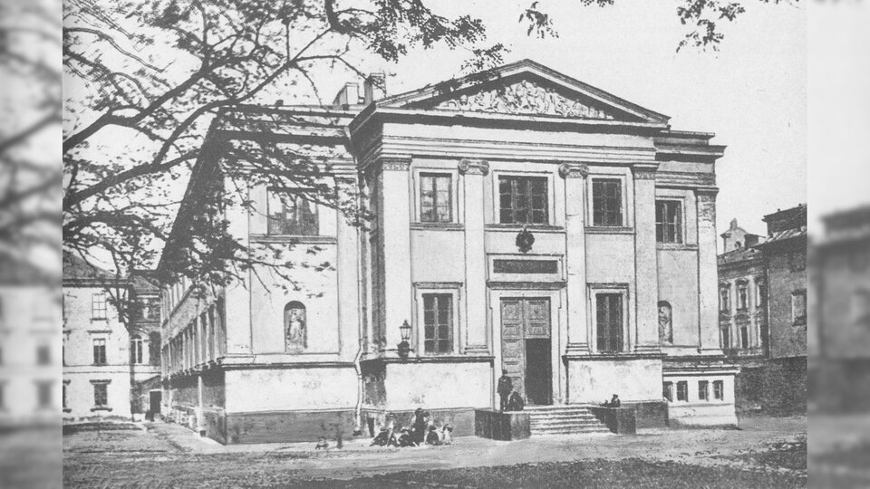 Obraz przedstawia duży, piętrowy budynek zbudowany w stylu neoklasycystycznym. Do budynku prowadzą schody.