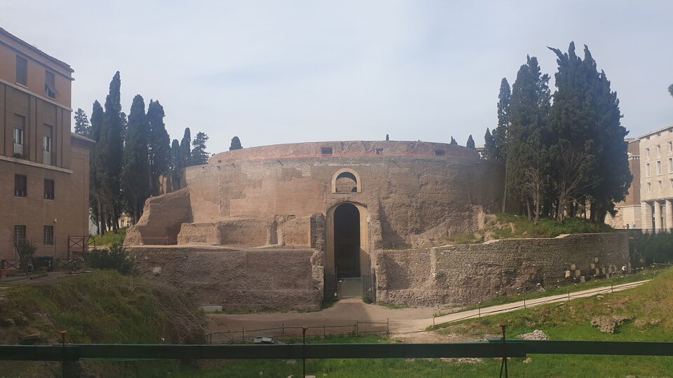 Mauzoleum Augusta w Rzymie - rotundowa budowla, z wąskim otworem wejściowym, otoczona niewysokim murem;  po prawej i lewej stronie budowli wysokie ciemnozielone drzewa.
