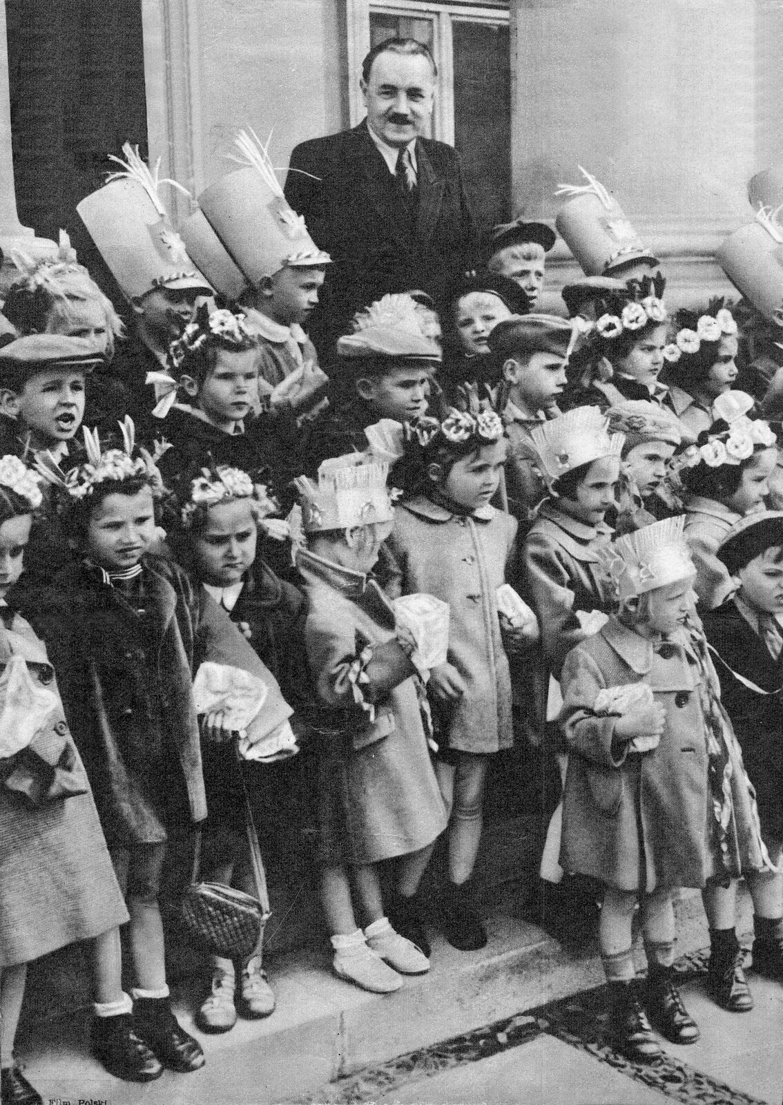 Mężczyzna ubrany w garnitur stoi w ostatnim szeregu z dziećmi. Stoją na schodach, w tle znajduje się ściana budynku i okno. Dziewczynki na głowach mają wianki. Chłopcy noszą czapki z kartonu imitujące wojskowe nakrycia głowy.