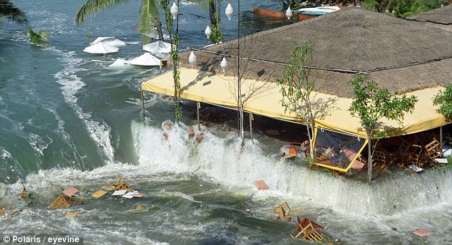 Zdjęcie przedstawia tsunami w Tajlandii. Na pierwszym planie w wodzie pływają krzesła i inne meble. Dalej jest budynek przez który przelewa się woda. W jego środku znajdują się porozrzucane meble. W tle w wodzie pływają parasole.