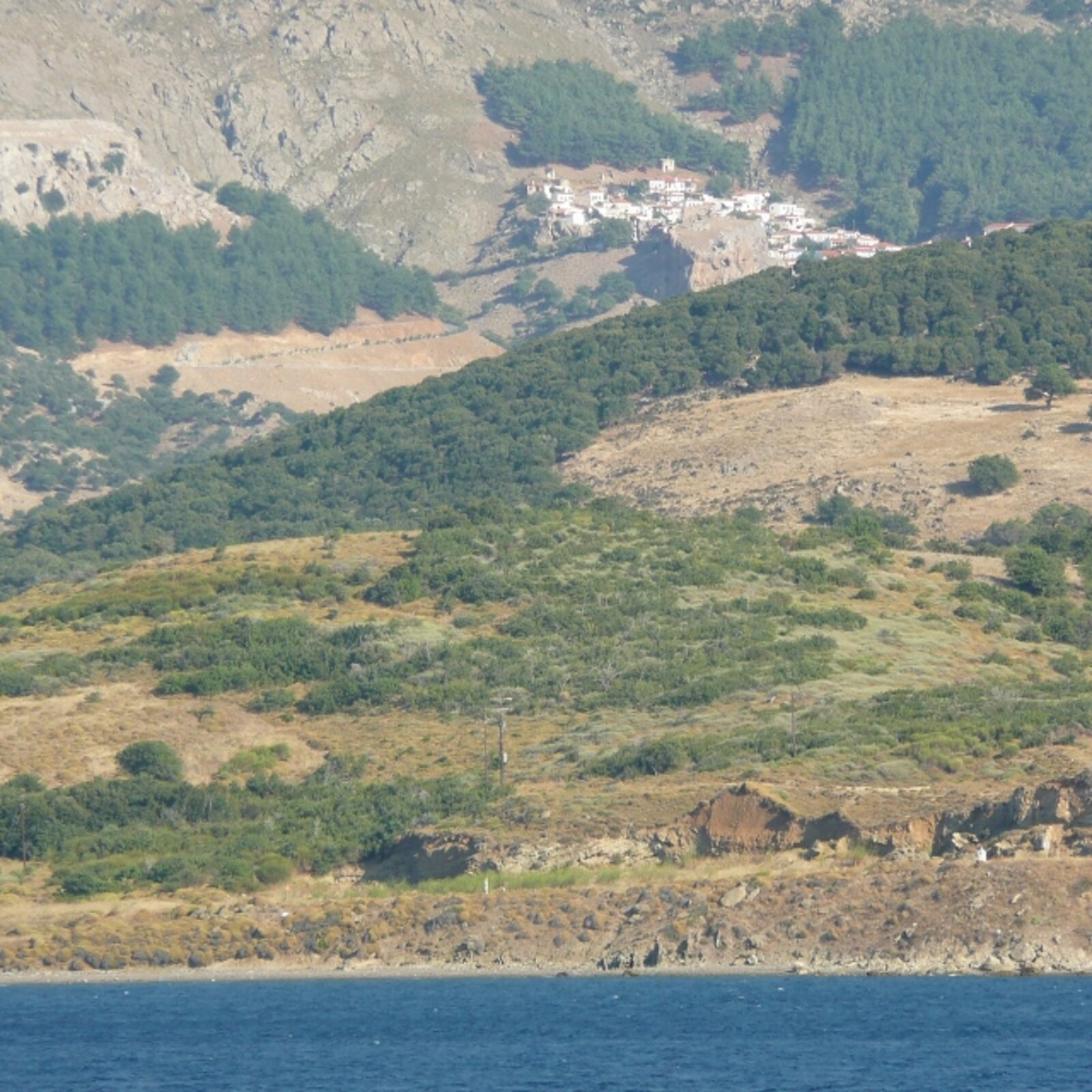 Ilustracja przedstawia kolorową fotografią greckiego miasteczka Kamariotissa Samothrakis. Na pierwszym planie ukazana jest błękitna tafla wody. Za nią znajdują się wzgórza porośnięte zaroślami. Tuż za nimi, u podnóża wysokich skalistych gór, znajdują się nieliczne białe zabudowania z czerwonymi dachami.