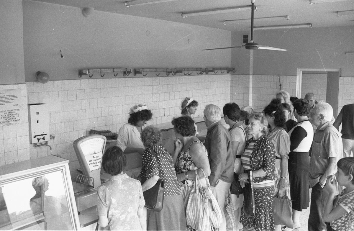 Zdjęcie przedstawia wnętrze sklepu mięsnego, w którym nie ma żadnego towaru. Za ladą stoją dwie sprzedawczynie w fartuchach. Za nimi goła ściana w kafelkach. Na ladzie waga szalkowa. Obok niej oszklona gablota. Przed ladą tłoczą się ludzie. Stoją i czekają na towar, którego na razie nie ma.