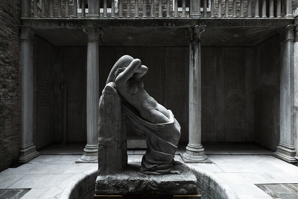 Na zdjęciu widzimy posąg nagiej kobiety, która górną częścią pleców opiera się o niski mur. Odzienie, które miała na sobie opadło jej prawie do kolan. Widać nagie piersi i brzuch. Kobieta ma długie włosy i zasłania twarz skrzyżowanymi rękami. W tle znajduje się fragment balustrady wsparty na kamiennych kolumnach.