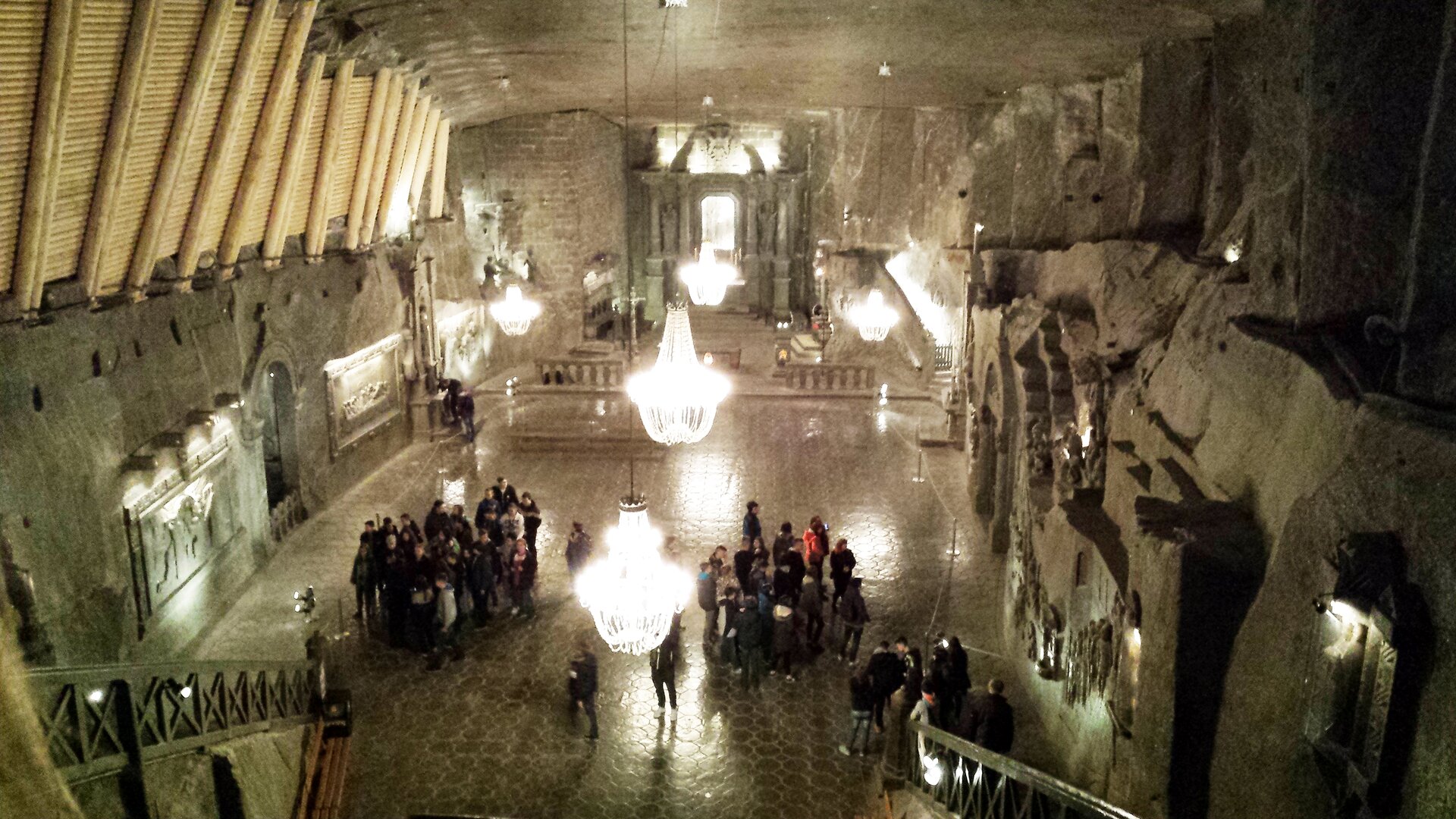 Ilustracja przedstawia dwie grupy osób podczas zwiedzania kopalni soli. Ściany pomieszczenia oraz podłoga są wykonane z kamienia. Pod sufitem znajdują się liczne żyrandole.