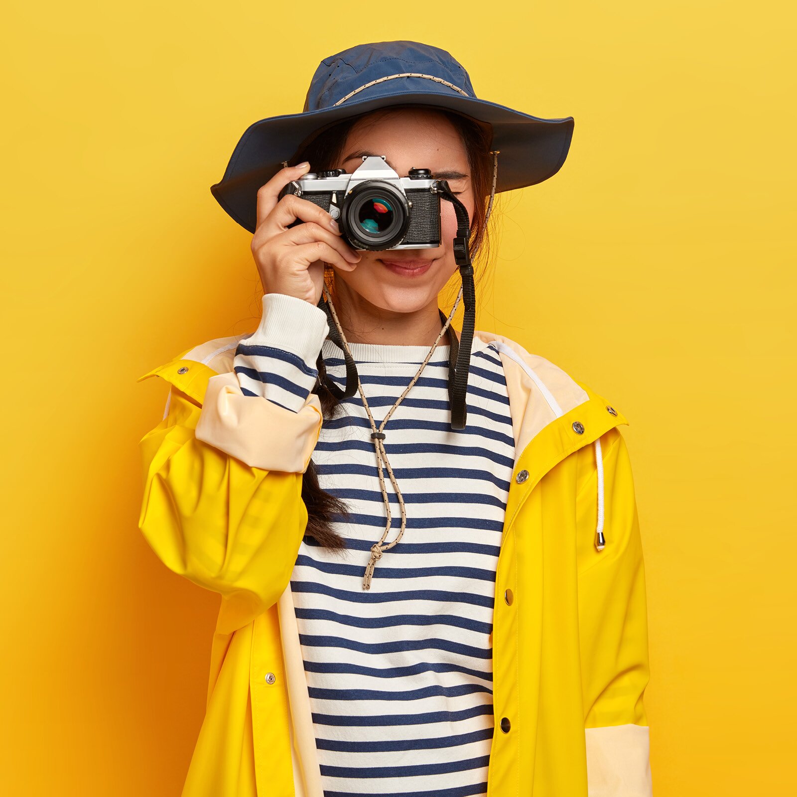 Zdjęcie przestawia dziewczynkę w wieku szkolnym, ubraną w niebieski kapelusz i żółtą kurtkę, patrzącą na nas przez obiektyw aparatu fotograficznego. 