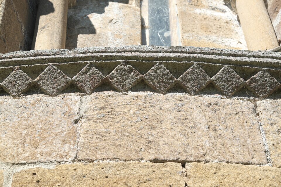 Fragment romańskiego muru z jasnego kamienia, z dekoracyjnym fryzem złożonym z rytmicznie powtarzających się rombów biegnących w poziomym pasie pod gzymsem.