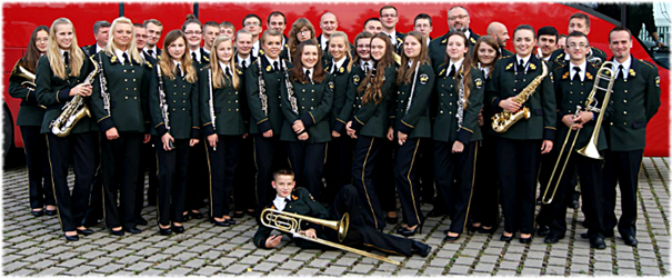 Zdjęcie przedstawia muzyków (dorosłych ludzi) w mundurach strażackich. Niektóre osoby trzymają w rękach instrumenty: saksofon, trąbkę. Przed nimi leży chłopiec, trzyma w ręku trąbkę. 