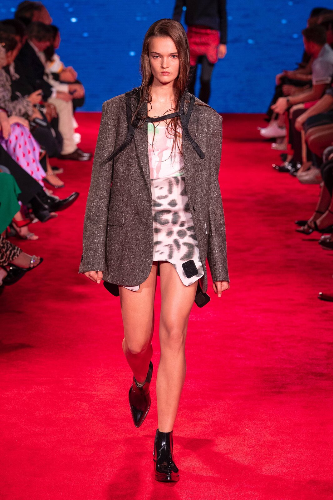 Fotografia przedstawia modelkę na wybiegu. Ma ona  na sobie krótką spódnicę w cętki, białą koszulkę z różowymi akcentami oraz obszerną, siwą marynarkę. Buty kobiety są czarne, a ich czubki spiczaste. W tle widać kolejną modelkę oraz siedzących ludzi. Dywan, po którym chodzą modelki, jest czerwony. 
