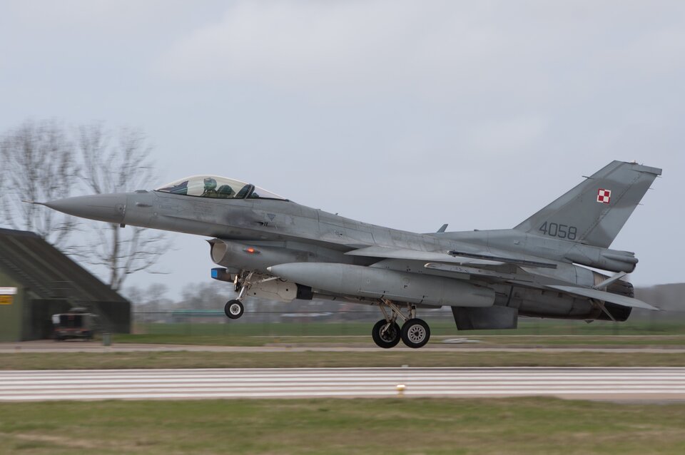 Fotografia przedstawia startujący samolot wojskowy. Dziób samolotu uniesiony w górę. Na sterze kierunku znajduje się biało‑czerwona szachownica. W tle budynki i bezlistne drzewa.