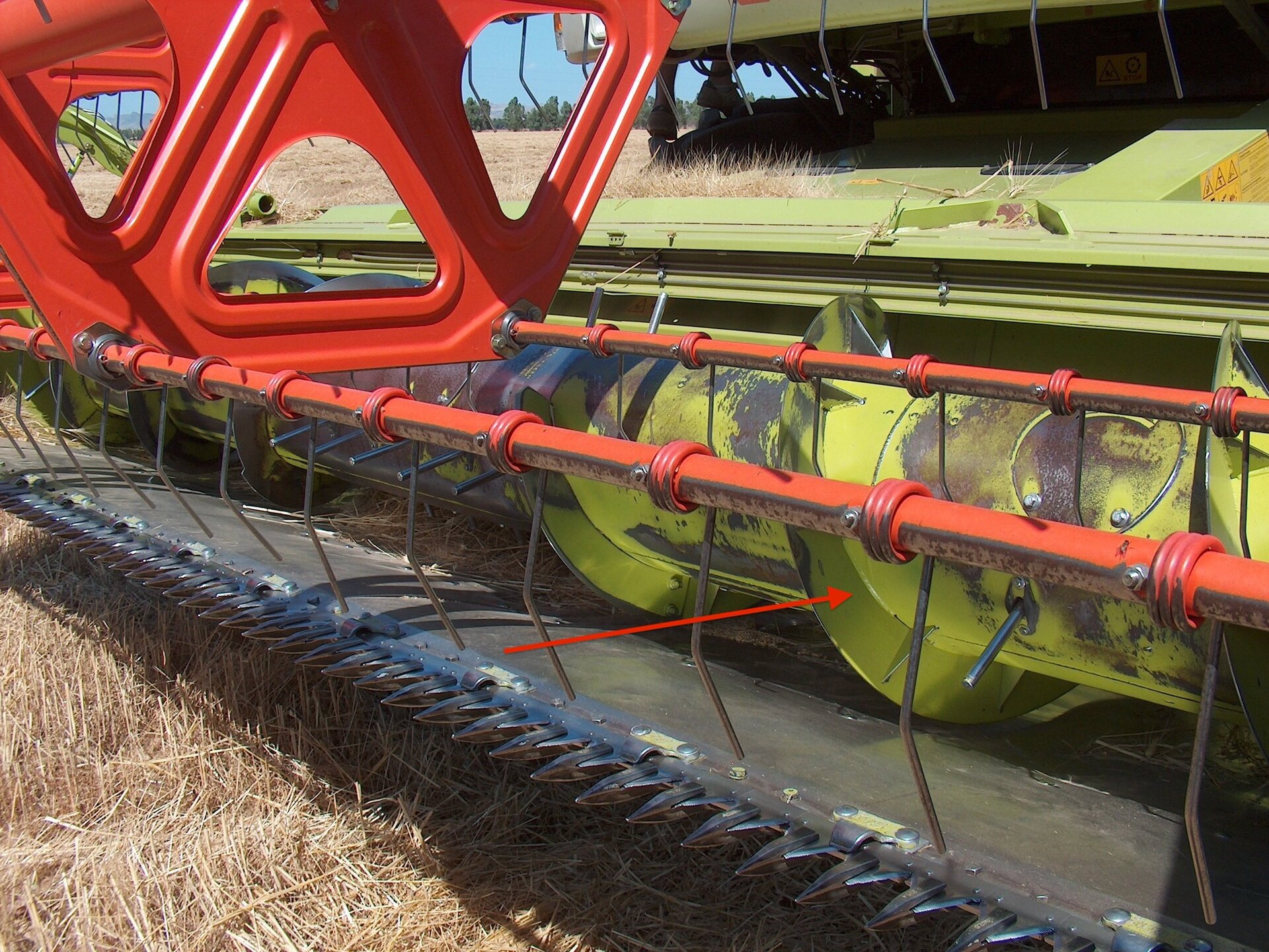 Zdjęcie przedstawia zbliżenie na heder kombajnu zbożowego. Czerwoną strzałką zaznaczono podajnik ślimakowo‑palcowy. Jest on osadzony na bokach obudowy zespołu żniwnego, za zespołem tnącym. Składa się z bębna, na który nawinięte są zwoje ślimakowe.