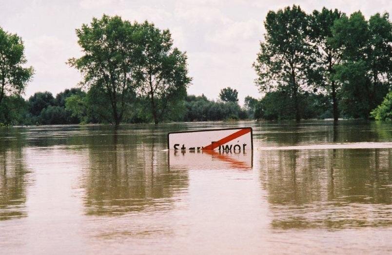 Zdjęcie przedstawia powódź w Starym Dworze. Woda sięga połowy wysokości tabliczki z nazwą miejscowości. W tle znajdują się zalane drzewa.