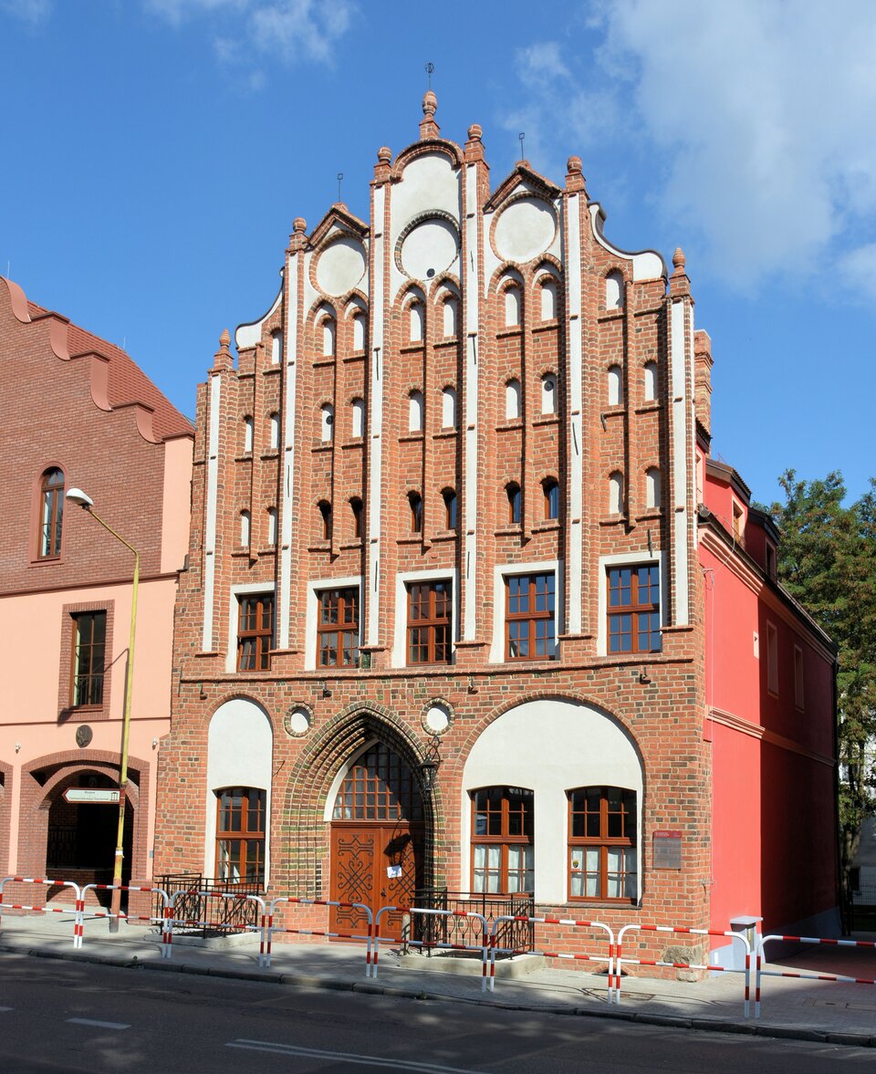 Wąska, ceglana kamienica w stylu gotyckim z dekoracyjnym, schodkowym szczytem i smukłymi, pionowymi blendami; na parterze łukowy portal i okna, przed budynkiem chodnik z barierkami, a nad nim jasne, częściowo zachmurzone niebo.