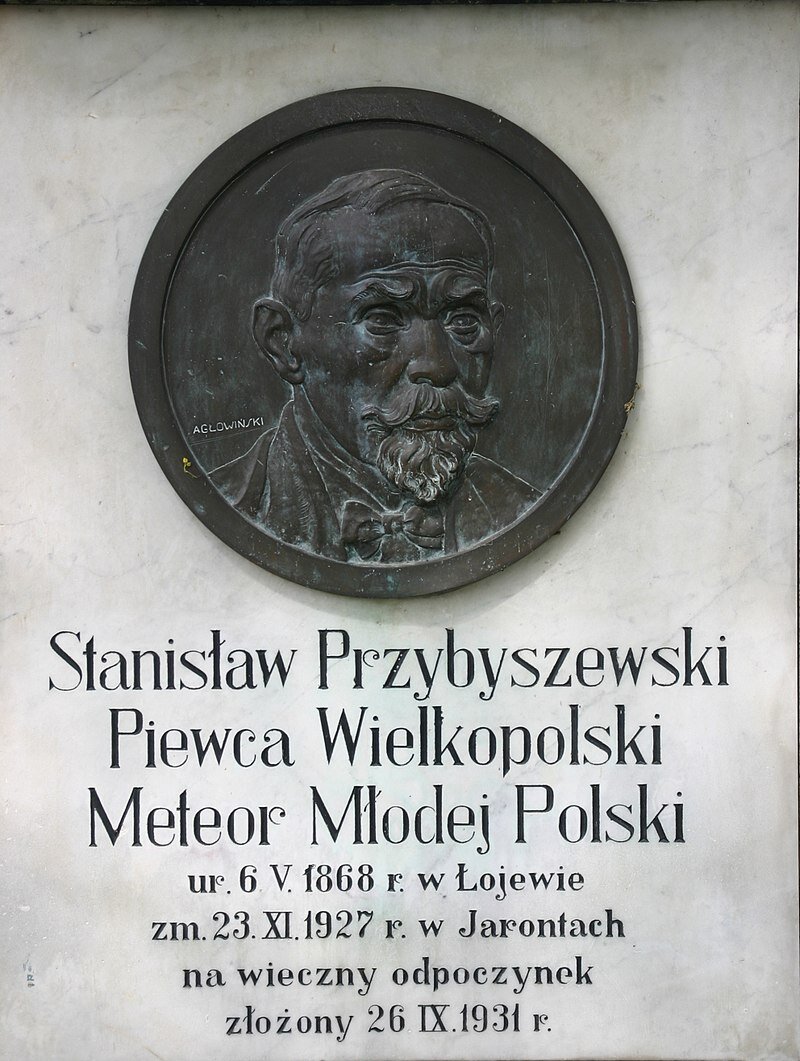 Ilustracja przedstawia tablicę nagrobną z wizerunkiem starszego mężczyzny o krótkich włosach, z brodą i wąsami, ubranego w marynarkę, koszulę i muszkę. Poniżej napis Stanisław Przybyszewski, Piewca Wielkopolski, Meteor Młodej Polski, urodzony 6 maja 1868 roku w Łojewie, zmarł 23 listopada 1927 roku w Jarontach, na wieczny odpoczynek złożony 26 września 1931 roku. 