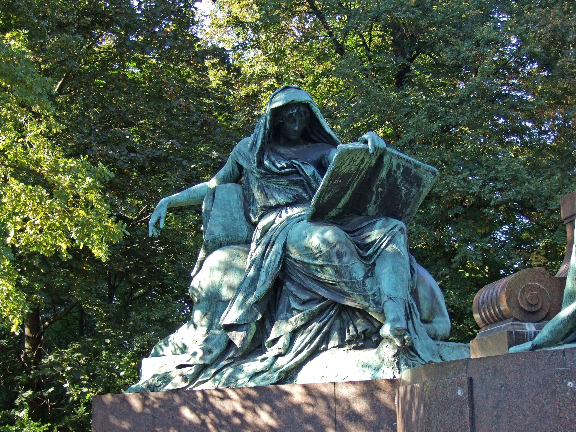 Ilustracja przedstawia fotografię pomnika „Sybilla”. Rzeźba jest w kolorze zielono- czarnym, a podstawa jest brązowa. Przedstawia siedzącą postać. Ubrana jest w długie szaty.  Na głowie ma zarzuconą chustę. W lewej dłoni trzyma otwartą książkę. Prawa spoczywa na oparciu krzesła. W tle są zielone drzewa liściaste. 