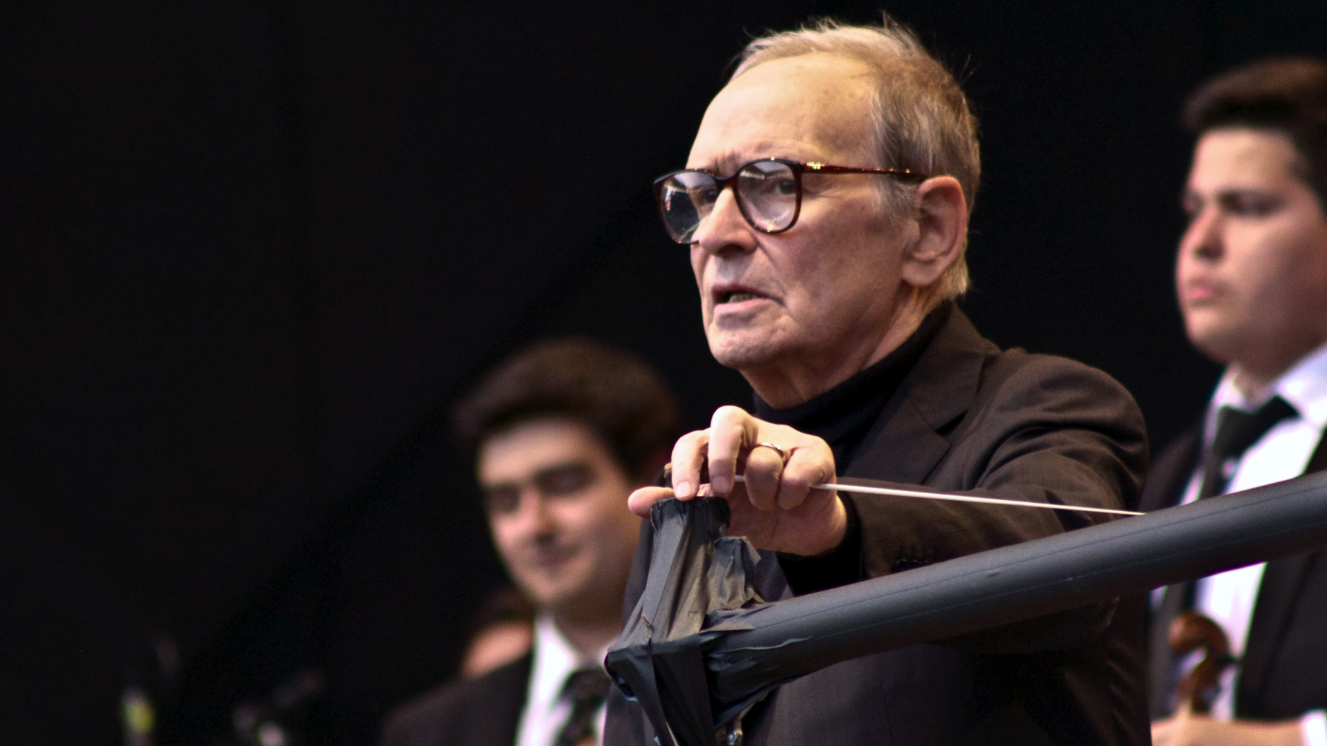 Zdjęcie przedstawia słynnego włoskiego kompozytora. W ręku trzyma batutę. Na drugim planie widać dwóch członków orkiestry.