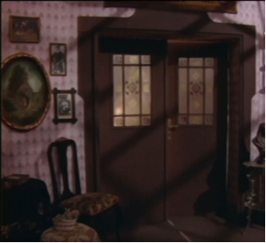 Zdjęcie przedstawia drewniane drzwi z okienkami. Przy drzwiach stoi krzesło, a na różowej tapecie wiszą obrazy.