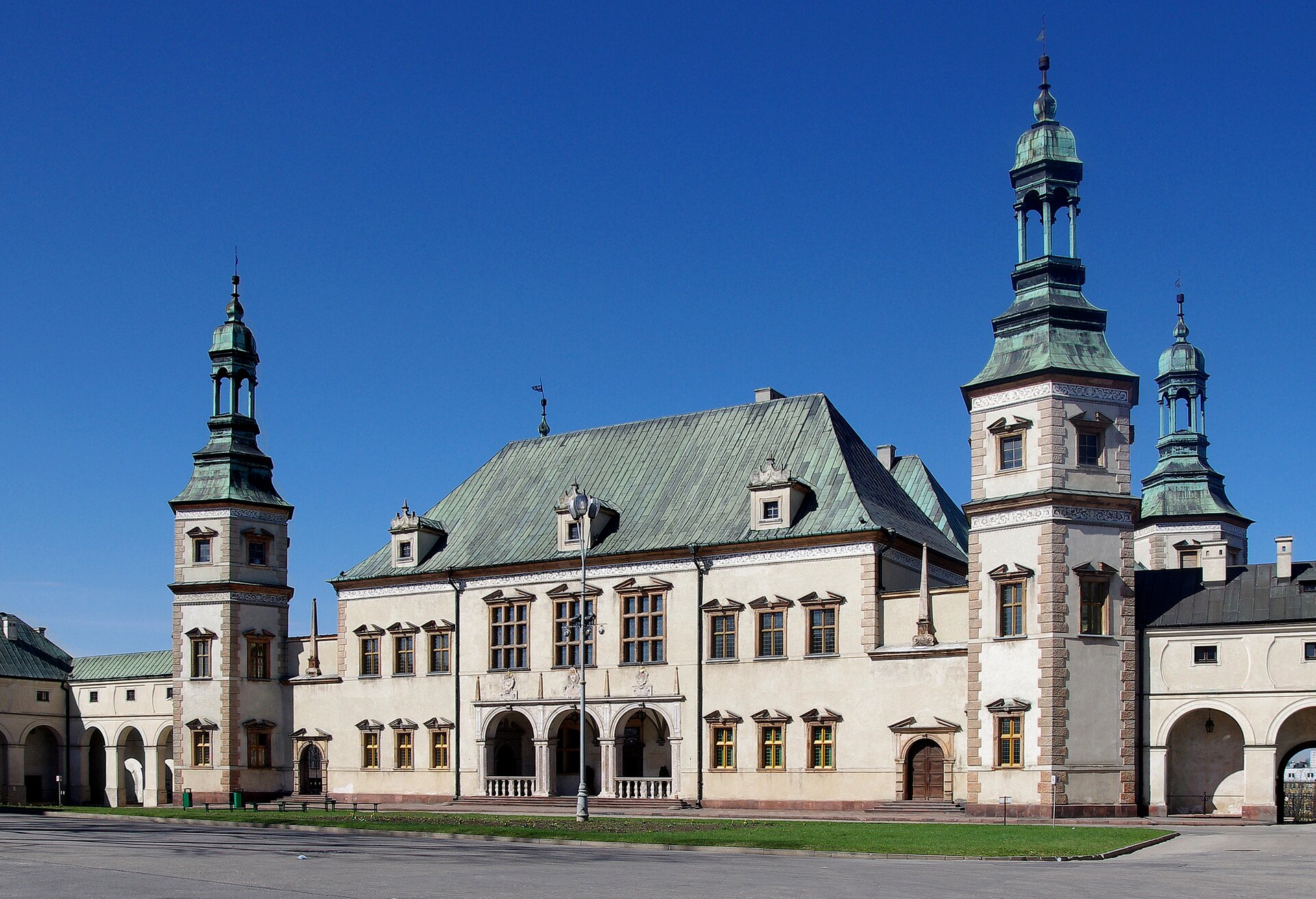 Zdjęcie przedstawia budynek pałacowy. Zbudowany został z jasno -  różowej cegły. Dach budynku pokrywa zielona dachówka. Centralnej części znajduje się wejście ozdobione trzema łukami. Po bokach znajdują się dwie, niewysokie wieże. Przed zamkiem znajduje się plac i trawniki. 