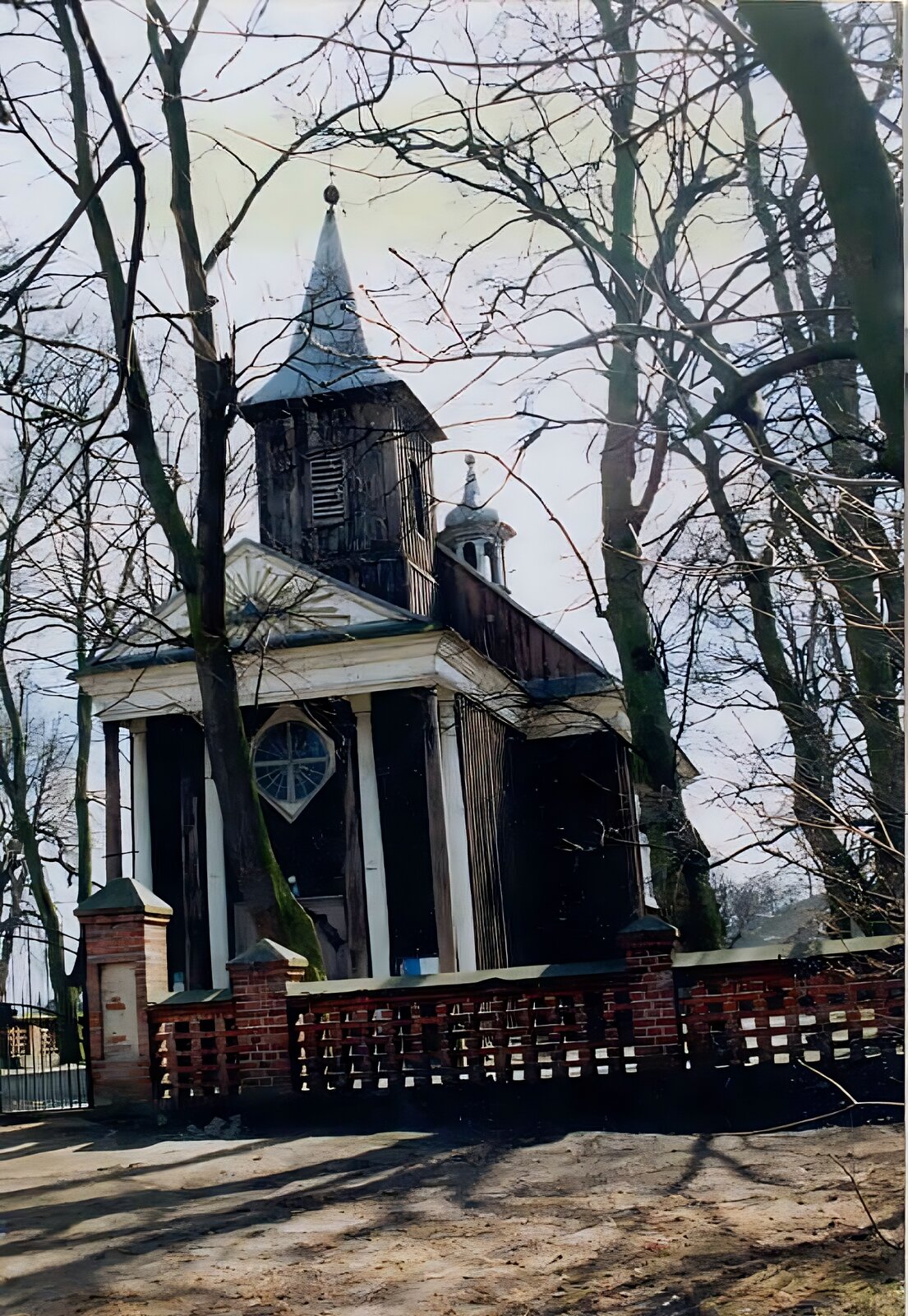 Zdjęcie przedstawia drewniany kościół, który ma skośny dach. Przy wejściu do kościoła znajdują się białe, wysokie kolumny. Nad wejściem znajduje się wieżyczka z szarym, spadzistym dachem. Teren kościoła został ogrodzony murowanym płotem wykonanym z czerwonej cegły. Wokół kościoła rosną wysokie drzewa, które nie mają liści. Zdjęcie zostało wykonane w świetle dziennym.