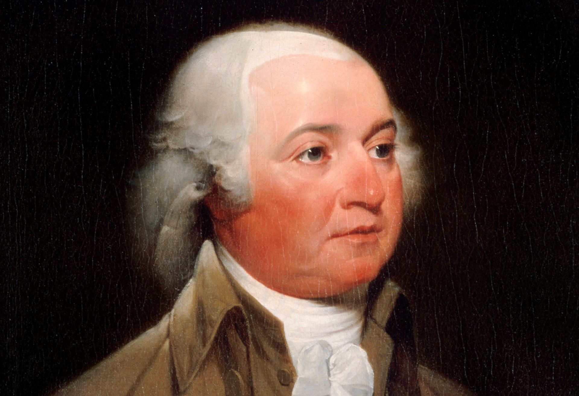 Zdjęcie przedstawia portret Johna Adamsa. Widoczna głowa mężczyzny w średnim wieku, zwrócona w prawo. Widoczna jest pełna twarz, lekko czerwona, duże, szeroko otwarte oczy, wydatny nos, usta, mocno zarysowany podbródek. Włosy mężczyzny są siwe, z tyłu dłuższe, z przodu ma zakola. Szyja osłonięta wysoką białą stójką, na której widoczny jest duży kołnierz w kolorze brązowym.