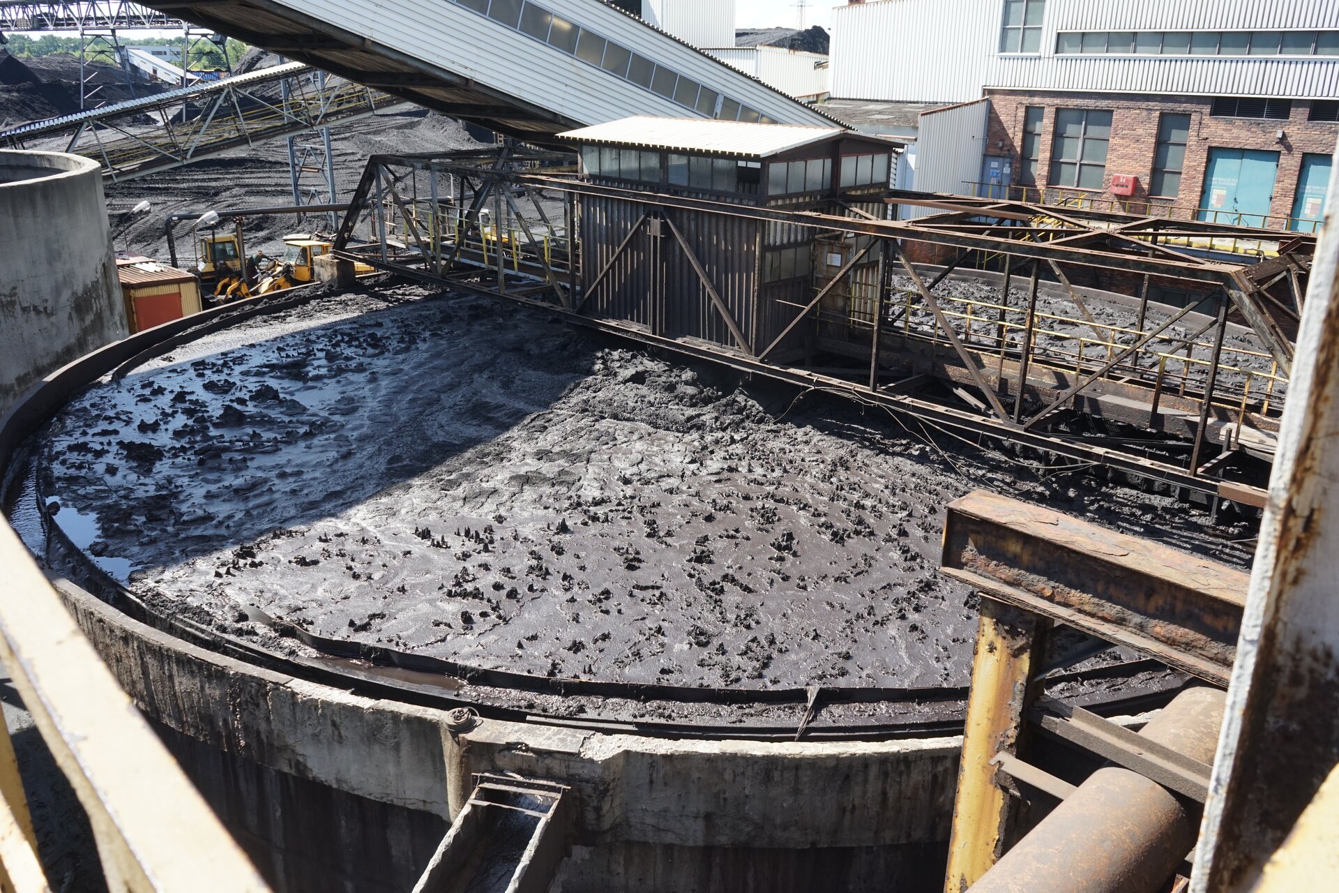 Zdjęcie przedstawia zagęszczacz promieniowy. Znajduje się on na otwartym powietrzu. Zagęszczacz zbudowany jest ze zbiornika na planie koła o dużej średnicy. Nad nim znajduje się pomost dla obsługi. Zanieczyszczona woda robocza doprowadzana jest rurociągiem do cylindrycznej rury nadawczej. Woda wypływa z niej do zagęszczacza poniżej zwierciadła wody i rozpływa się promieniowo w kierunku zewnętrznego obwodu. W miarę zmniejszania się prędkości przepływu na dno zagęszczacza opadają coraz to drobniejsze ziarna. Osiadające na dnie zbiornika ziarna zgarniane są obrotowym czteroramiennym zgarniaczem do wylotu stożkowego. Pompa mułowa odbiera zagęszczone ziarna z wylotu i przetłacza je do dalszych procesów przeróbczych. Oczyszczona woda przepływa do pierścieniowego koryta. skąd przelewem odprowadzana jest do rząpia pompowego. Poniżej zdjęcia znajduje się przycisk multimedialny z nagraniem audio. Treść nagrania jest zgodna z tytułem zdjęcia.