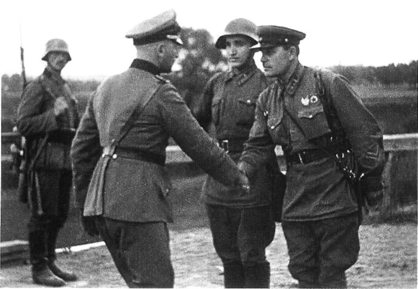 Czarno‑białe zdjęcie przedstawia czterech wojskowych. Dwóch z nich wita się przez uścisk dłoni. Trzeci stoi obok w hełmie na głowie, w oddali stoi czwarty żołnierz. Ma hełm na głowie, na ramieniu ma powieszony karabin. W tle las.   