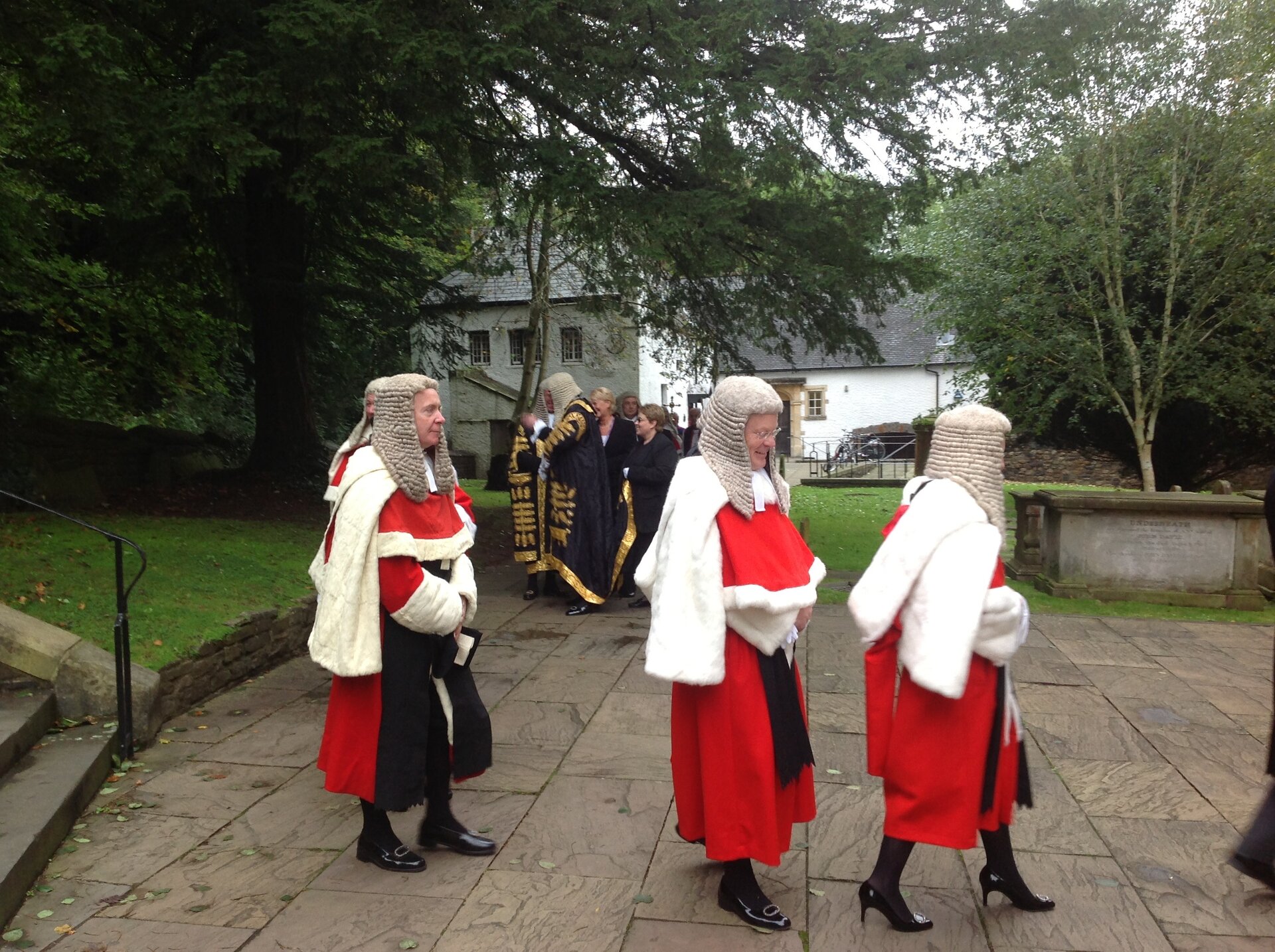Zdjęcie przedstawia idących chodnikiem sędziów. Są oni ubrani w czerwono-czarne togi z białymi dodatkami, na głowach mają długie peruki z włosami ułożonymi w loki.