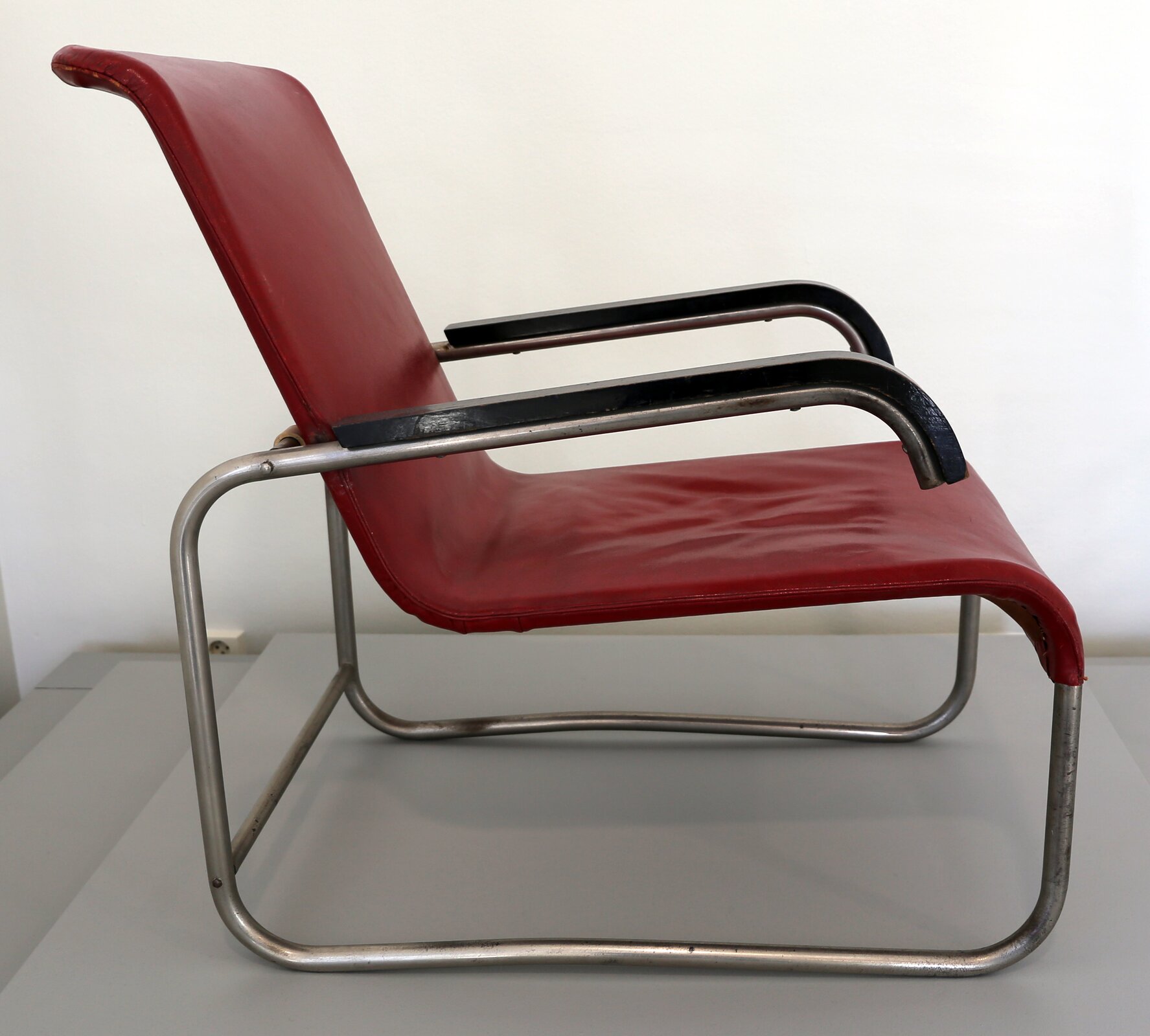 Praca Marcela Breuera pod tytułem „Krzesło B35”. W centrum zdjęcia stoi krzesło, którego konstrukcja została oparta na stalowych giętkich rurkach, do których przymocowano czerwone siedzisko wykonane ze skóry. Krzesło posiada dwie podpory wykonane z  czarnego materiału. Mebel postawiono na szarym podwyższeniu.