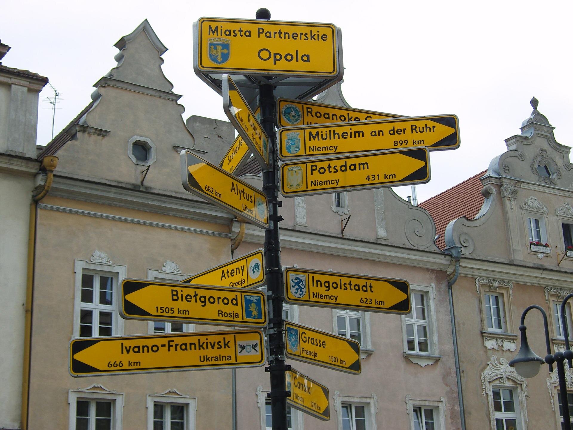 Tablice informujące o miastach bliźniaczych Opola. Na maszcie zamocowane są poziome żółte tabliczki z nazwami miast i państw, w których te miasta leżą. Każda z tabliczek zakończona jest strzałką i skierowana została w kierunku odpowiadającego jej miasta.