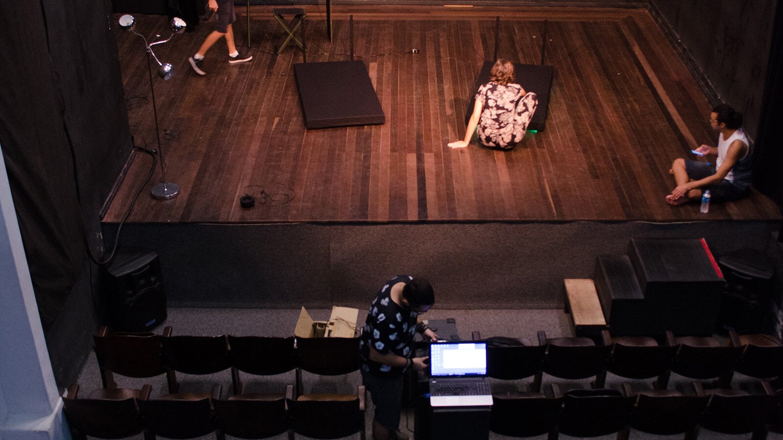 Zdjęcie przedstawia scenę w teatrze, na której ustawiono lampę i dwie czarne platformy. Na środku sceny widoczna jest kobieta kucająca przed jedną z platform. Z boku siedzi mężczyzna po turecku i patrzy w ekran telefonu. Przed sceną widoczny jest mężczyzna, który wpatrzony jest w ekran komputera.