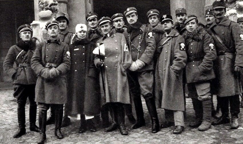 Fotografia przedstawia przedstawia grupę żołnierzy stojących przed budynkiem. Na prawym ramieniu mają naszywki ze znakiem ludzkiej czaszki. Wśród nich jest jedna kobieta, która ma na głowie chustkę ze znakiem krzyża.