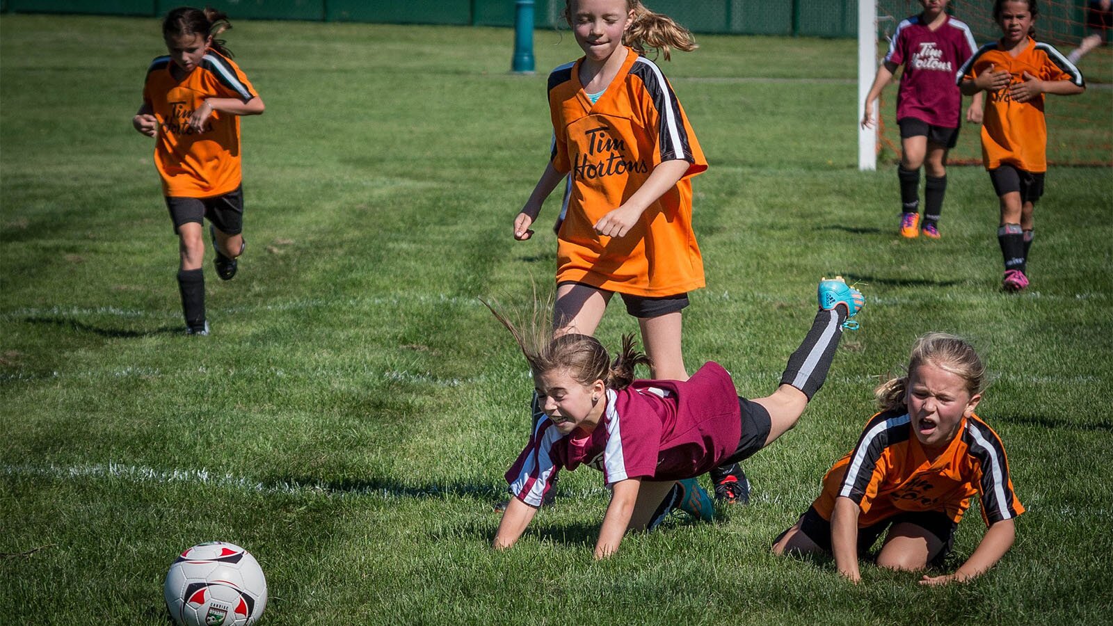 Zdjęcie przedstawia dwie drużyny dziewcząt podczas meczu w piłkę nożną. Dziewczynki ubrane są w stroje piłkarskie. Jedna drużyna ma pomarańczowe koszulki, a druga bordowe. Na pierwszym planie widoczne są dwie dziewczynki leżące na murawie. W tle widoczne są biegnące dziewczynki.